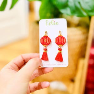 May include: Red lantern earrings with gold detailing and red tassels, displayed on a white card. The card features the name "Eflie" in green script and a website address. The earrings are a festive accessory.