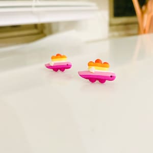 May include: A pair of small, pink, yellow, orange, and white dinosaur earrings. The dinosaurs are facing the right side of the image.