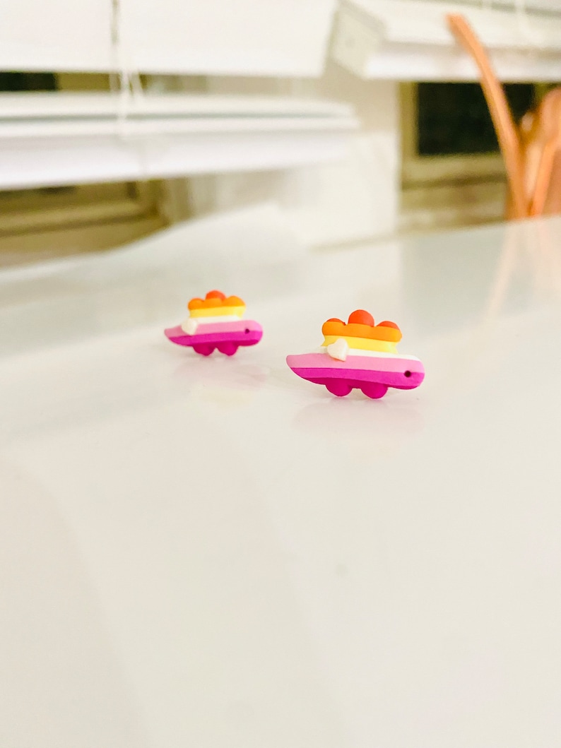 May include: A pair of small, pink, yellow, orange, and white dinosaur earrings. The dinosaurs are facing the right side of the image.