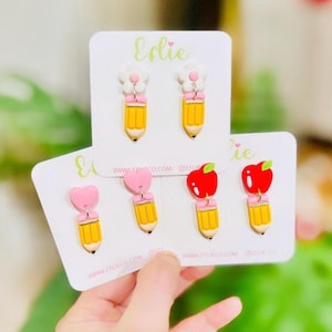 May include: Three sets of earrings featuring a yellow pencil charm with a different topper. The first set has white flower toppers, the second set has pink heart toppers, and the third set has red apple toppers. The earrings are displayed on white cards with the text "Ellie" and "www.ellieco.com".