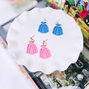 May include: Four pairs of handmade earrings shaped like princess dresses. Two pairs are light blue, and two are pink. The earrings are displayed on a white, ruffled plate. The earrings have silver and gold-colored ear wires.