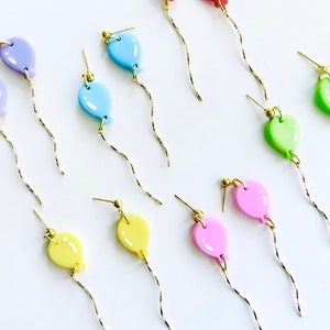 Puede incluir: Un conjunto de pendientes colgantes de globos de colores. Los pendientes están hechos de arcilla polimérica y presentan una variedad de colores, incluyendo rojo, rosa, amarillo, verde, azul y morado. Cada pendiente tiene una cadena dorada que cuelga del globo.