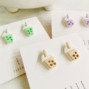 May include: A set of three pairs of handmade polymer clay earrings. Each pair features a boba tea cup with different colored drinks and toppings. The colors are green, purple, and brown.