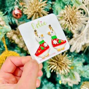 Op de afbeelding: Kerst-thema oorbellen met witte ganzenfiguren die rode truien dragen met groene accenten en sneeuwvlok- en boomontwerpen. De oorbellen hebben goudkleurige haken en worden gepresenteerd op een witte kaart met de tekst "Eflie" en een websiteadres.