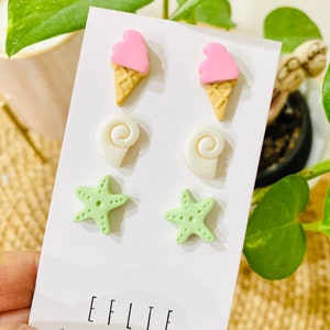 May include: A set of six colorful polymer clay earrings. The earrings are shaped like ice cream cones, seashells, and starfish. The ice cream cones are pink and white, the seashells are white, and the starfish are green. The earrings are displayed on a white card with the text "EFLIE HANDMADE WITH LOVE".