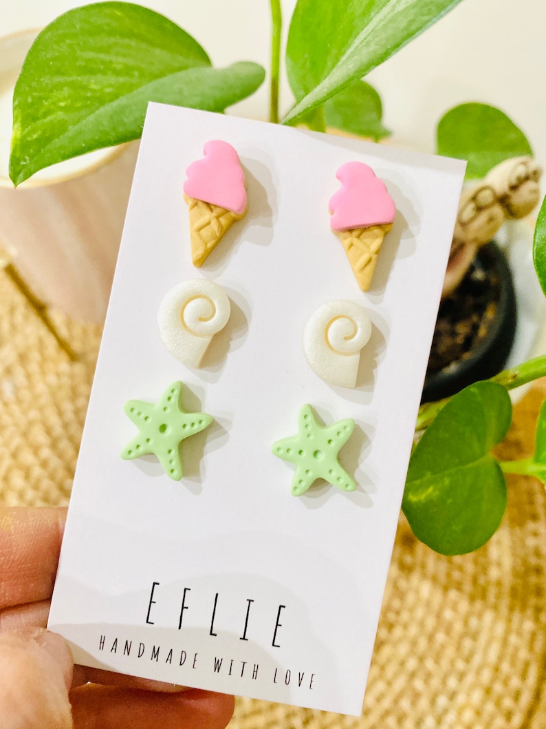 May include: A set of six colorful polymer clay earrings. The earrings are shaped like ice cream cones, seashells, and starfish. The ice cream cones are pink and white, the seashells are white, and the starfish are green. The earrings are displayed on a white card with the text "EFLIE HANDMADE WITH LOVE".