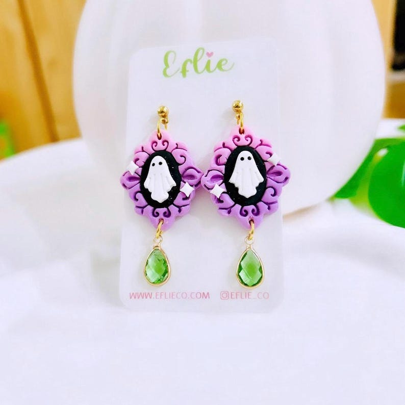 May include: A pair of Halloween-themed earrings. Each earring features a purple and pink frame with a white ghost design in the center. Green teardrop-shaped gems dangle below. The earrings are displayed on a white card with the text "Eflie".