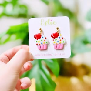 May include: A pair of cupcake earrings with a pink base, white frosting, colorful sprinkles, and a red heart on top. The earrings are on a white card with the word "Elie" and the website address "www.eflieco.com".