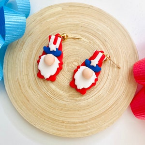 May include: A pair of red, white, and blue gnome earrings with gold hooks. The gnomes are wearing a red, white, and blue hat with a star on top.