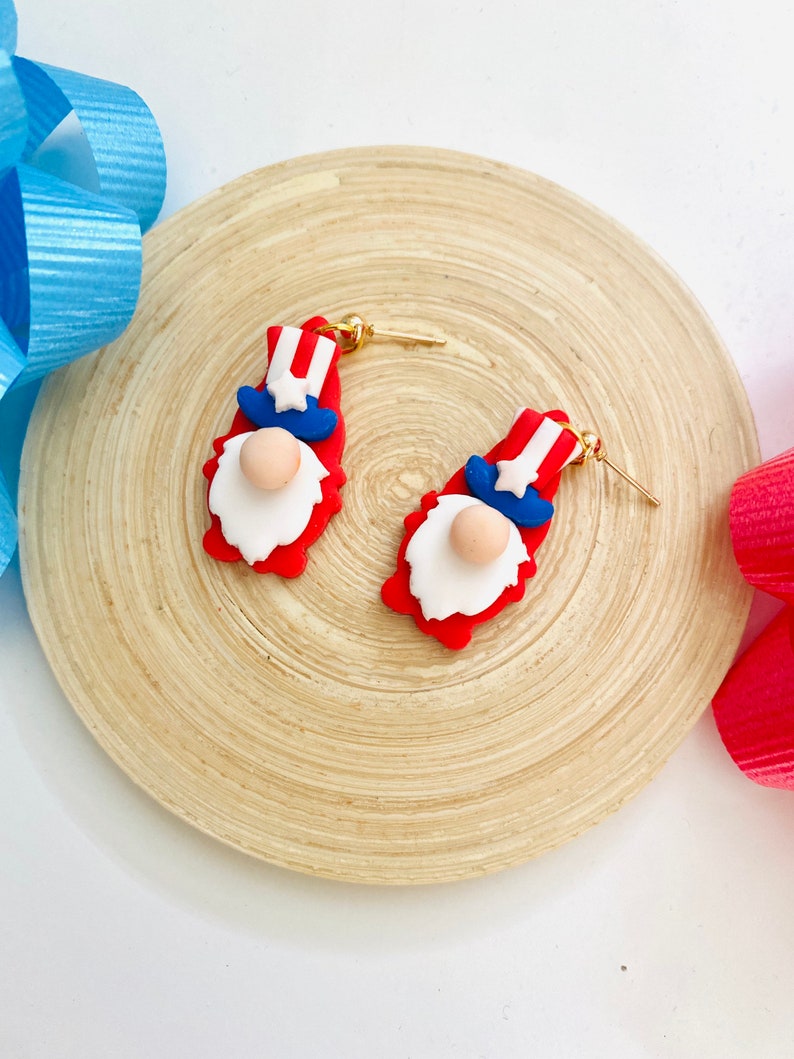 May include: A pair of red, white, and blue gnome earrings with gold hooks. The gnomes are wearing a red, white, and blue hat with a star on top.