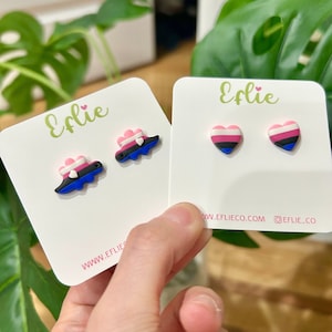 May include: Two pairs of stud earrings on white cards. One pair features a cloud design with pink, white, blue, and black accents. The other pair is heart-shaped, with pink, white, blue, and black stripes. The cards have the text "Eflie" and website information.