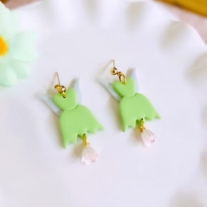 May include: A pair of whimsical earrings featuring a fairy design. Each earring has a light green dress shape with gold-colored accents and a small pink flower charm. The earrings have a light blue wing design.