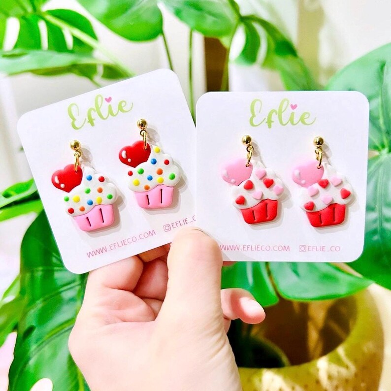 May include: Two pairs of cupcake earrings. One pair is pink and white with red hearts. The other pair is white and red with rainbow sprinkles. Both pairs have gold hooks.