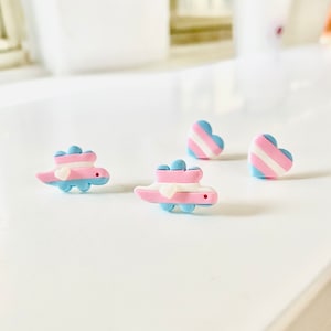 May include: Three pairs of earrings featuring a transgender pride flag design. Two pairs are heart-shaped and one pair is a small dinosaur. The earrings are made of polymer clay and are painted in shades of blue, pink, and white.