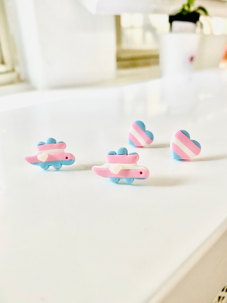 May include: Three pairs of earrings featuring a transgender pride flag design. Two pairs are heart-shaped and one pair is a small dinosaur. The earrings are made of polymer clay and are painted in shades of blue, pink, and white.