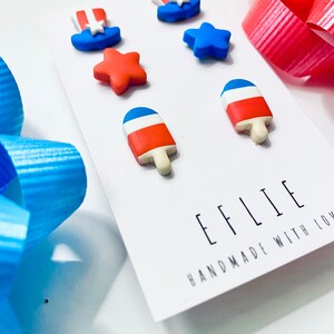 May include: A set of six red, white, and blue polymer clay earrings. The earrings are shaped like stars, popsicles, and top hats. The earrings are handmade and have the text "EFFLIE HANDMADE WITH LOVE" on the card.