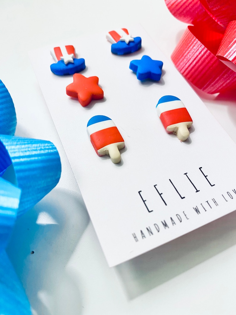May include: A set of six red, white, and blue polymer clay earrings. The earrings are shaped like stars, popsicles, and top hats. The earrings are handmade and have the text "EFFLIE HANDMADE WITH LOVE" on the card.
