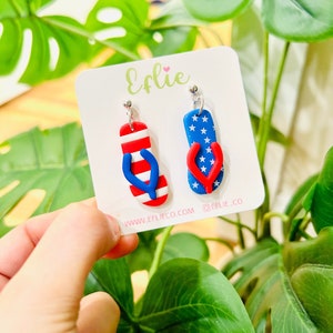 May include: A pair of red, white, and blue flip-flop earrings. The earrings are made of polymer clay and have a silver hook. The earrings are on a white card with the text "Effie" and "www.effieco.com @effie.co".