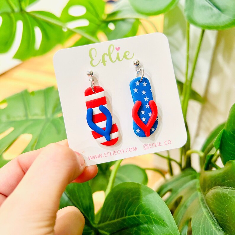 May include: A pair of red, white, and blue flip-flop earrings. The earrings are made of polymer clay and have a silver hook. The earrings are on a white card with the text "Effie" and "www.effieco.com @effie.co".