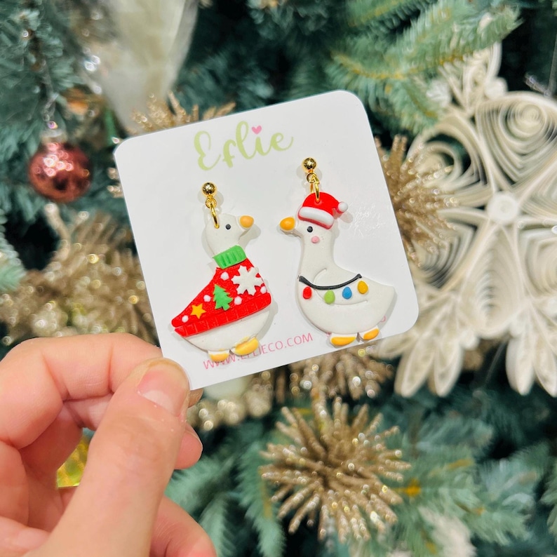 Op de afbeelding: Een paar kerstoorbellen met witte ganzenontwerpen. E&eacute;n gans draagt een rode trui met een groene kerstboom, de andere heeft een kerstmuts en lichtslingers. De oorbellen zitten op een witte kaart met de tekst "Eflie" en "www.eflie.co.com".