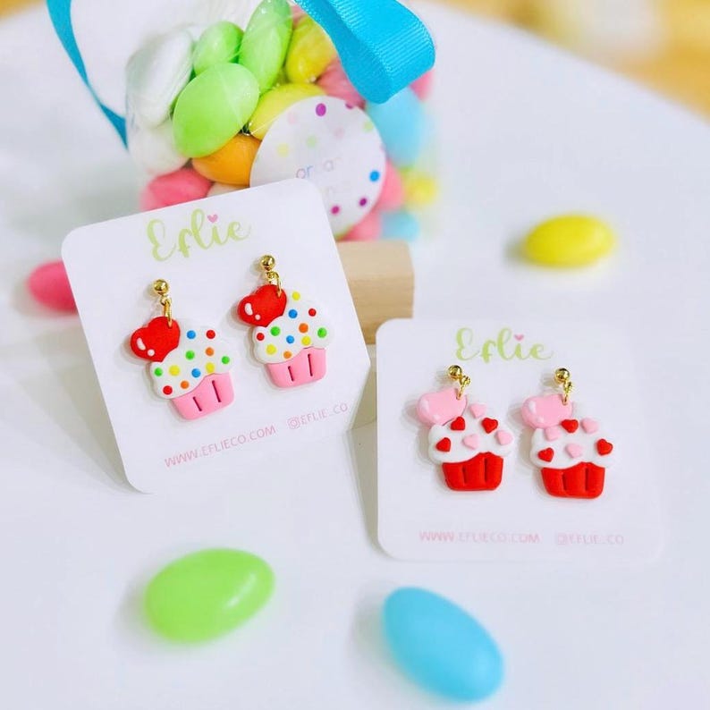 May include: Two pairs of cupcake earrings on white cards with the text "Edie" and website information. The earrings are made of polymer clay and feature colorful sprinkles and hearts. One pair is pink and white with red hearts, and the other pair is red and white with pink hearts.