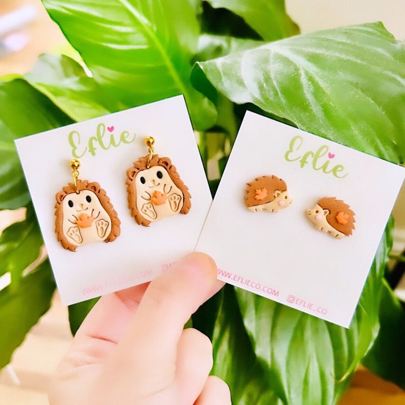 May include: Two pairs of hedgehog earrings displayed on white cards with the text "Elie" on them. The earrings are made from polymer clay and feature a hedgehog holding a leaf. The earrings are available to purchase on the website www.eflieco.com.