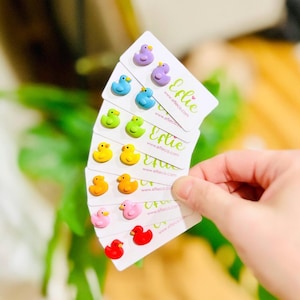 Può includere: Un set di 10 paia di orecchini a forma di papera di gomma colorati su carte bianche con il testo "Ellie" e "www.ellieco.com".