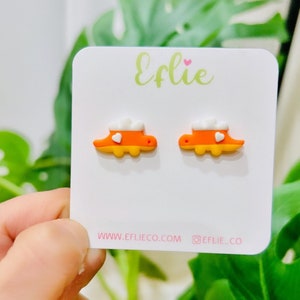 K&ouml;nnte beinhalten: Ein Paar orangefarbene und gelbe Ohrstecker in Dinosaurierform mit wei&szlig;en wolkenartigen Akzenten und einem kleinen wei&szlig;en Herz. Die Ohrringe werden auf einer wei&szlig;en Karte mit dem Text "Eflie" und der Website-Adresse www.eflieco.com pr&auml;sentiert.