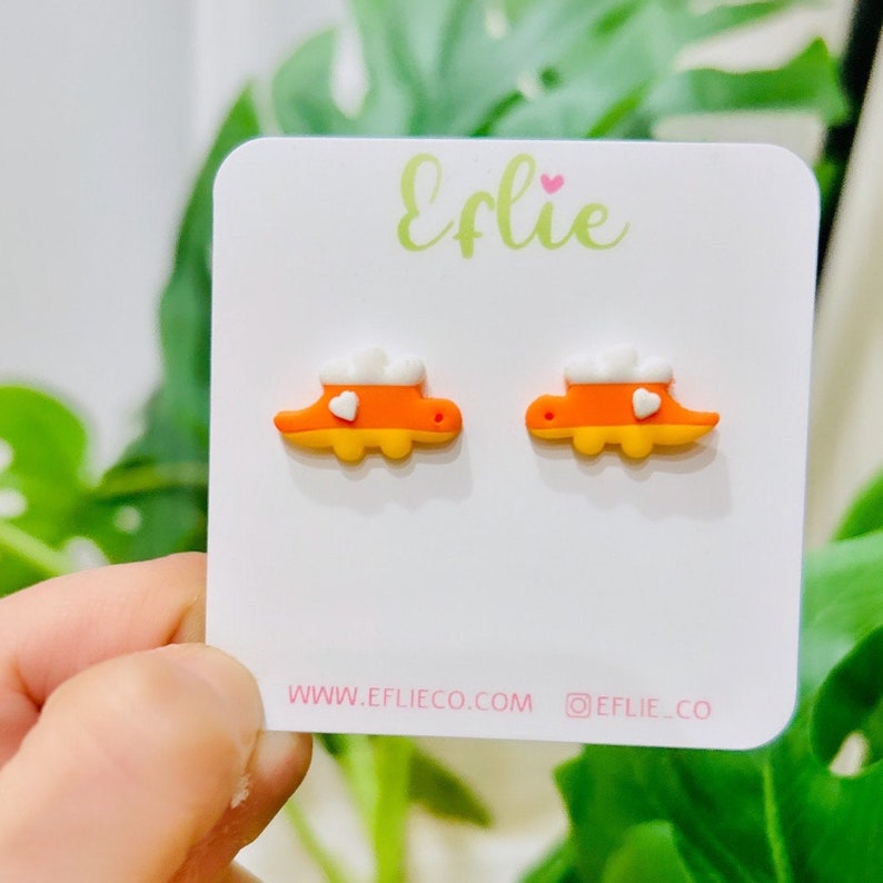 K&ouml;nnte beinhalten: Ein Paar orangefarbene und gelbe Ohrstecker in Dinosaurierform mit wei&szlig;en wolkenartigen Akzenten und einem kleinen wei&szlig;en Herz. Die Ohrringe werden auf einer wei&szlig;en Karte mit dem Text "Eflie" und der Website-Adresse www.eflieco.com pr&auml;sentiert.