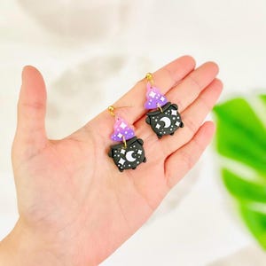 May include: A pair of black and purple witch cauldron earrings with gold-colored hooks. Each earring features a cauldron design with a crescent moon and star details, topped with a purple witch hat with white star accents. The earrings are held in a hand.
