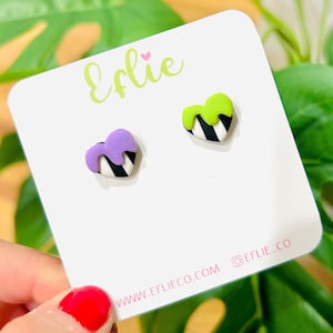May include: A pair of heart-shaped earrings with black and white stripes and a drip of purple and green glaze. The earrings are displayed on a white card with the text "Eflie" and "www.eflieco.com @eflie_co".