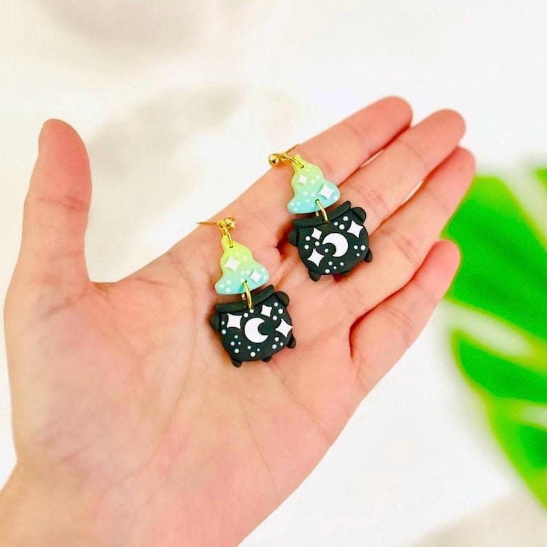May include: A pair of whimsical earrings featuring a witch's cauldron design. The earrings are black with white stars and a crescent moon. Above the cauldron is a witch hat in shades of green and blue. The earrings are attached to gold-colored ear wires.