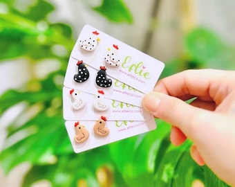 May include: Four pairs of polymer clay chicken earrings on white cards. The earrings are in white, black and white polka dot, brown, and white. The cards have the website address "www.eflieco.com" and the word "Elie" printed on them.