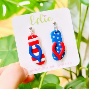 May include: A pair of red, white, and blue flip-flop earrings. The flip-flops are made of polymer clay and have a star pattern on one side and red and white stripes on the other. The earrings are on a white card with the text "Eflie" and "www.eflieco.com @eflie_co".