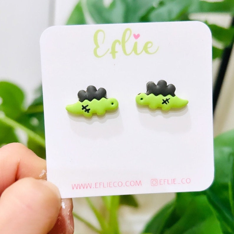 K&ouml;nnte beinhalten: Ein Paar gr&uuml;ne Dinosaurier-Ohrstecker mit schwarzen Stacheln und gestickten Details. Die Ohrringe werden auf einer wei&szlig;en Karte mit dem Markennamen "Eflie" in gr&uuml;ner Schrift pr&auml;sentiert. Die Karte enth&auml;lt auch die Website-Adresse und den Social-Media-Handle.