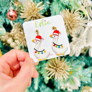 Op de afbeelding: Een paar feestelijke oorbellen met witte ganzenontwerpen met rode kerstmutsen en kleurrijke kerstverlichting. De oorbellen worden gepresenteerd op een witte kaart met de naam "Erlie" in groene letters.