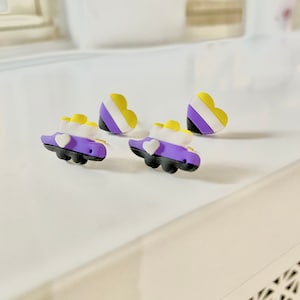May include: Non-binary pride earrings. The earrings feature a purple car shape with a white heart and yellow accents. Two heart-shaped charms with yellow, white, and purple stripes are also present. The earrings are on a white surface.