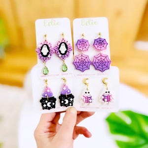 May include: A collection of Halloween-themed earrings. Designs include ghosts, spiderwebs, witch hats, and ghosts with books. The earrings are in shades of purple, pink, black, and white, with gold-colored hardware and green teardrop accents.