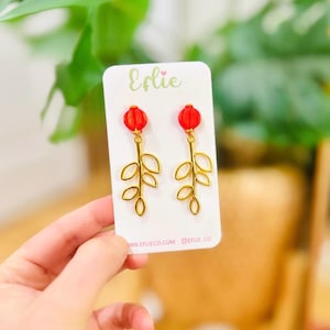 May include: Gold-coloured leaf-shaped earrings with red, round tops. The earrings are presented on a white card with the brand name "Eflie" in cursive. The card also includes the website address.