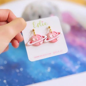 Op de afbeelding: Een paar roze, planeetvormige bungelende oorbellen met witte ringen en kleine steraccenten, bevestigd aan een witte displaykaart met de tekst Eflie, www.eflieco.com en @eflie.co. De oorbellen hebben goudkleurige metalen stekers.