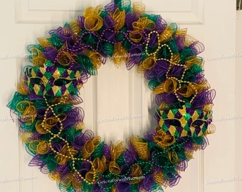 Puede incluir: Una corona de Mardi Gras decorada con malla morada, verde y dorada, cuentas doradas y dos máscaras de carnaval doradas y verdes.