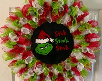 Könnte beinhalten: Ein Weihnachtskranz, der mit rotem, weißem und grünem Netz sowie silbernen und goldenen Ornamenten verziert ist. Der Kranz zeigt einen schwarzen Kreis mit einer grünen Grinch-Figur, die einen Weihnachtsmann-Hut trägt, und dem Text "Stink. Stank. Stunk."