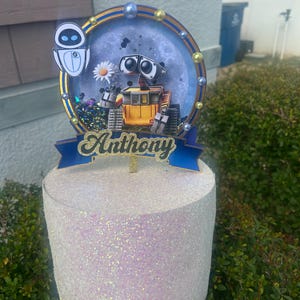May include: A cake topper featuring the characters Wall-E and Eve, with the name "Anthony" in gold script. The topper has a blue and gold circular frame with a starry background and a white glittery base.