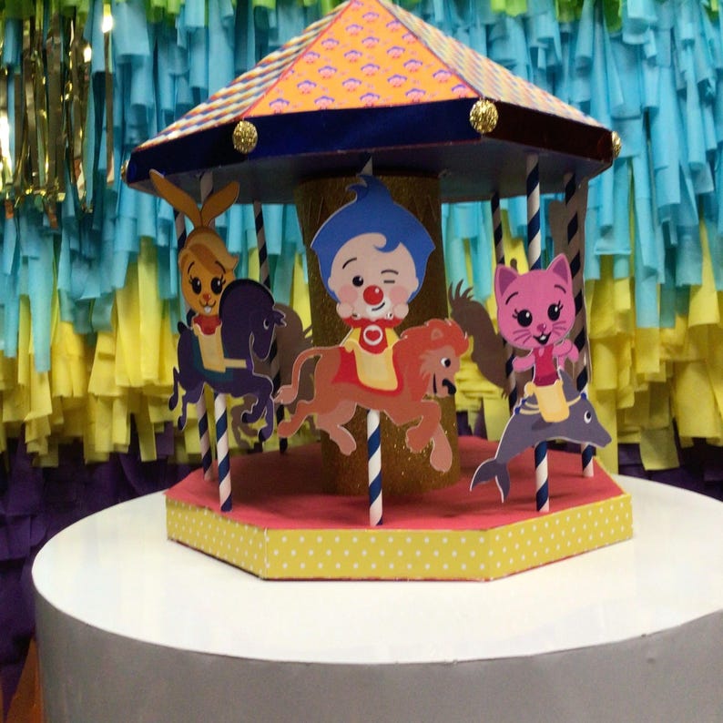 May include: A colorful carousel cake topper featuring cartoon characters riding animals. The carousel has a patterned roof and a yellow and white polka dot base. The characters include a bunny, a clown, and a pink cat.