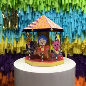 May include: A colorful carousel cake topper with cartoon characters riding animals. The carousel has a yellow and orange roof, and the base is yellow with white polka dots. The background is a colorful streamer backdrop.