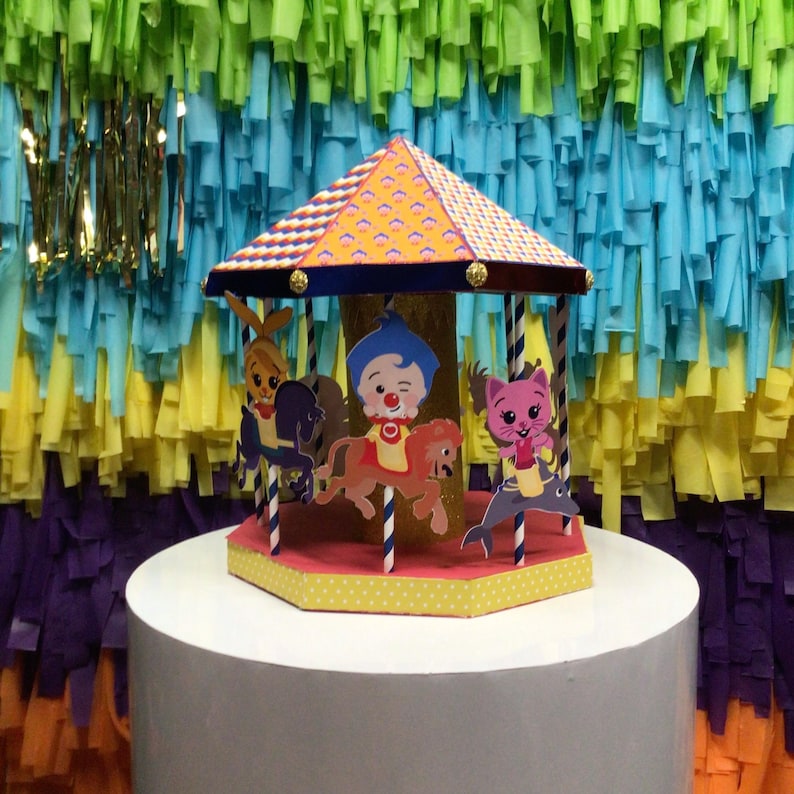 May include: A colorful carousel cake topper with cartoon characters riding animals. The carousel has a yellow and orange roof, and the base is yellow with white polka dots. The background is a colorful streamer backdrop.