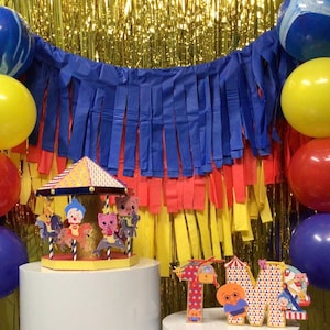 May include: A colorful carousel-themed party decoration with a yellow and orange roof, featuring cartoon characters. The scene includes blue, yellow, and red balloons, a blue and red fringe backdrop, and letter cutouts spelling "1M".