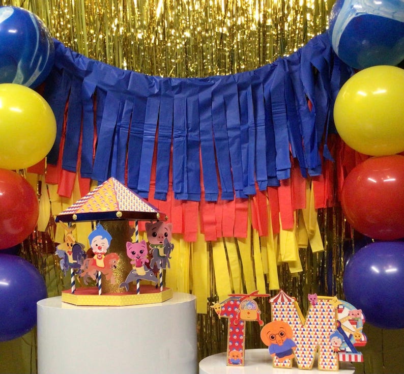 May include: A colorful carousel-themed party decoration with a yellow and orange roof, featuring cartoon characters. The scene includes blue, yellow, and red balloons, a blue and red fringe backdrop, and letter cutouts spelling "1M".