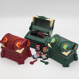 May include: Three decorative treasure chest-shaped game cartridge holders in red, green, and dark green. The chests have a stylized design with yellow and white accents. Several game cartridges and small red and green game pieces are visible.