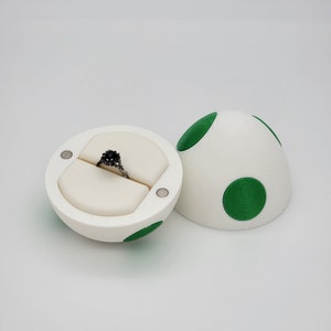May include: A white ring box shaped like an egg with green spots. The box is open and a black ring with a black stone is inside.